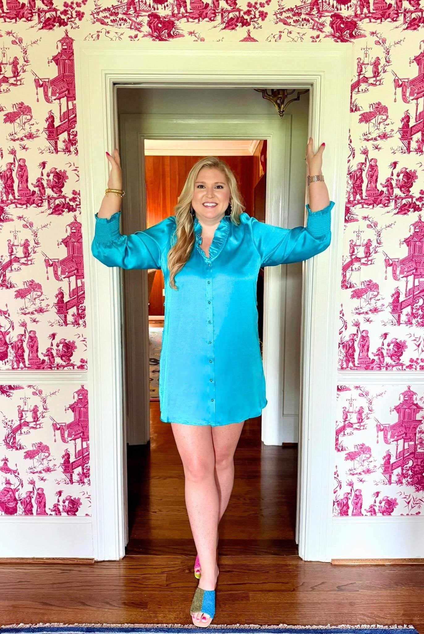 Woman wearing Harper Long Sleeve Dress in blue with ruffle neckline and gold buttons standing in doorway
