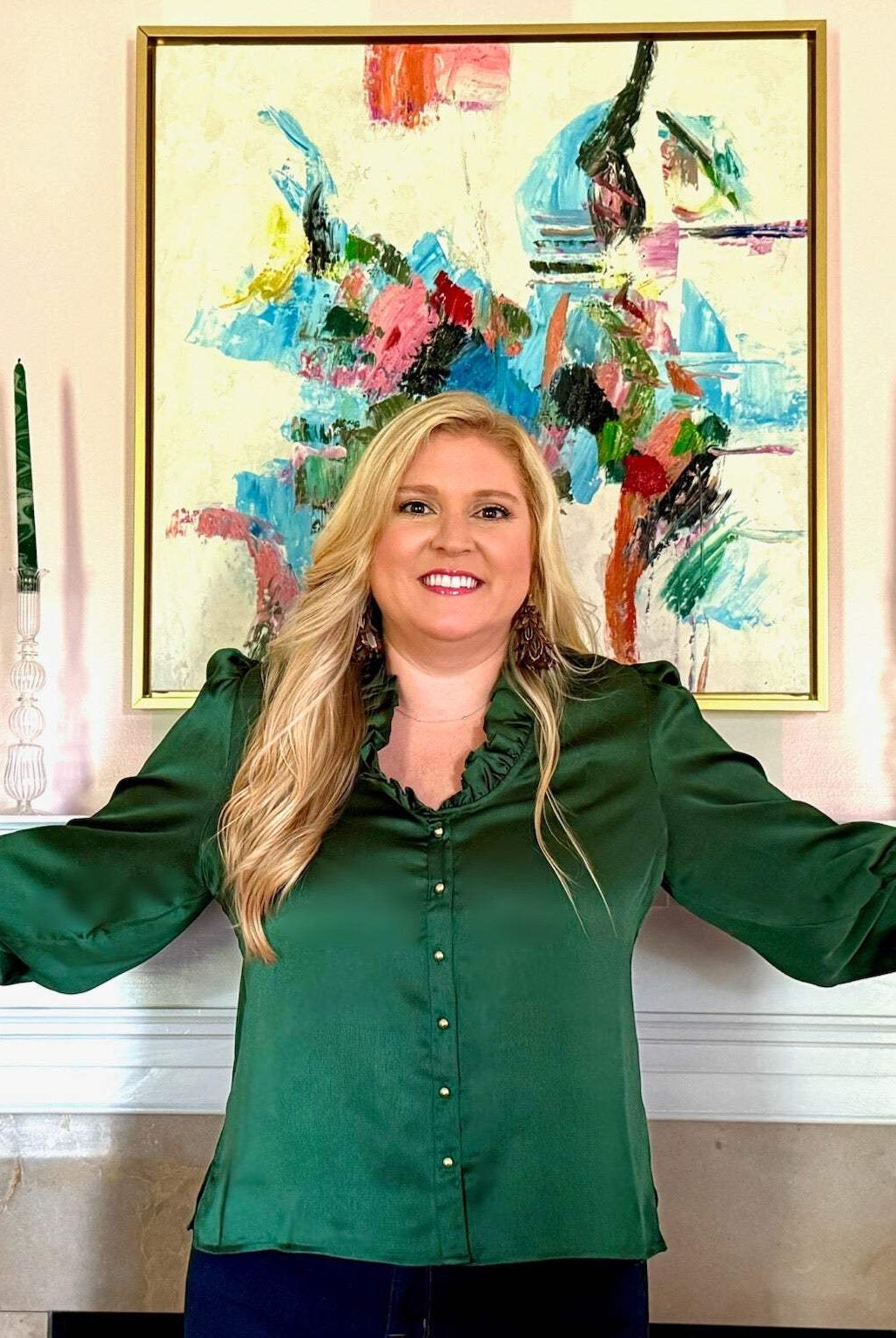 Woman wearing green Harper Top with ruffle neckline and gold buttons standing by mantelpiece with decorative plates and candles