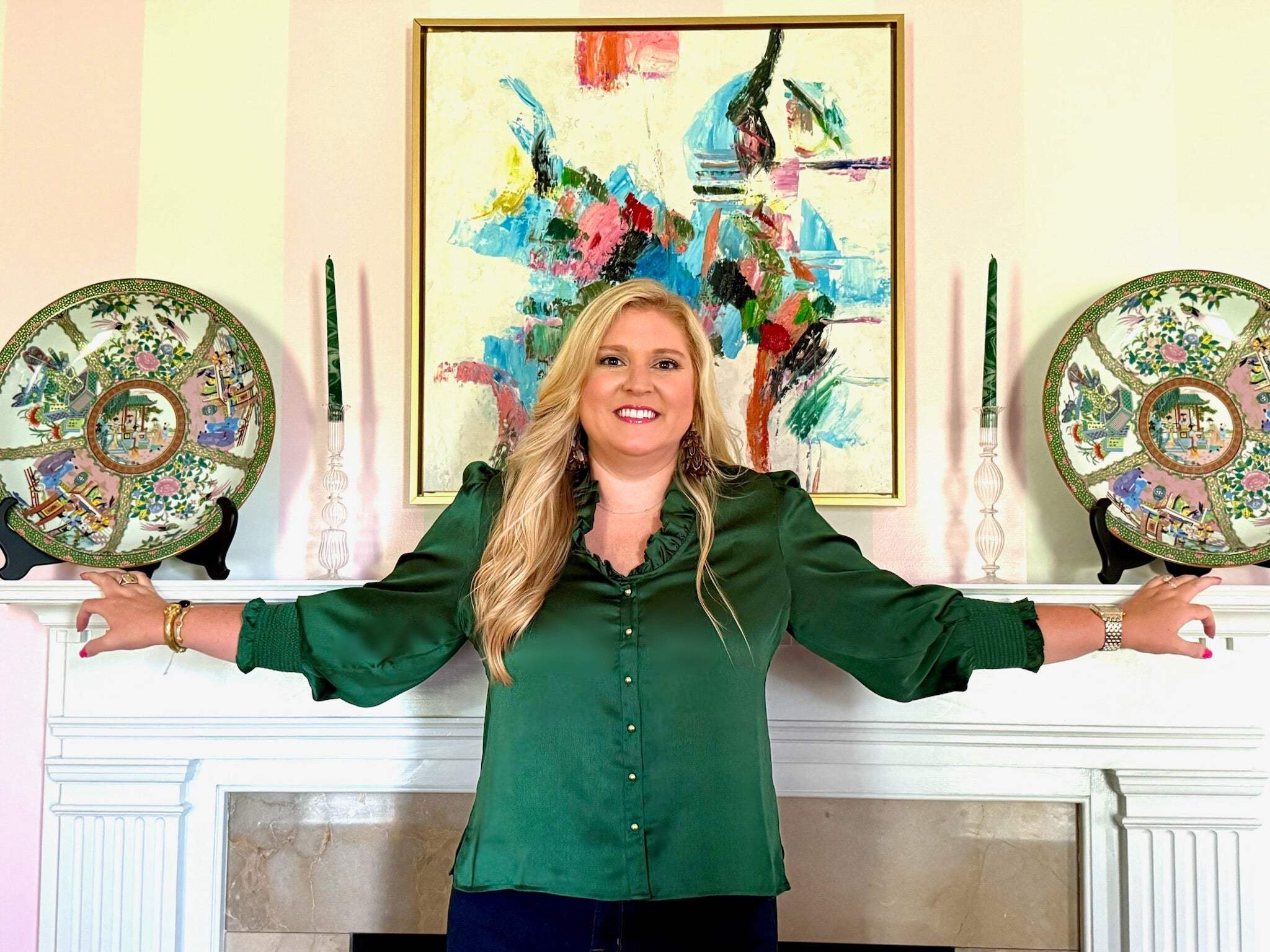 Woman wearing green Harper Top with ruffle neckline and gold buttons standing by mantelpiece with decorative plates and candles