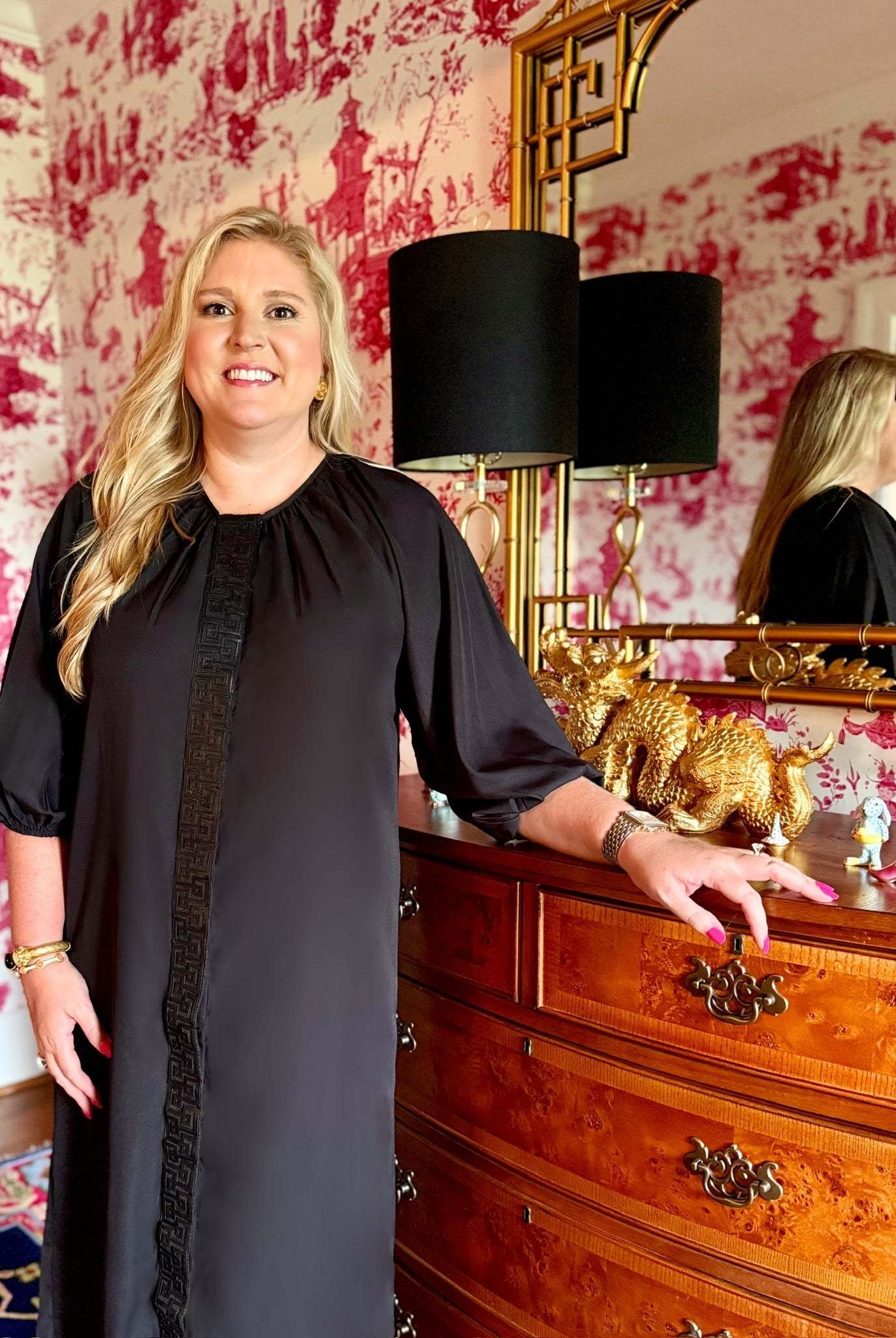 Woman wearing black Fall Hampton Dress with leather Greek key detail standing by wooden dresser with gold dragon decor