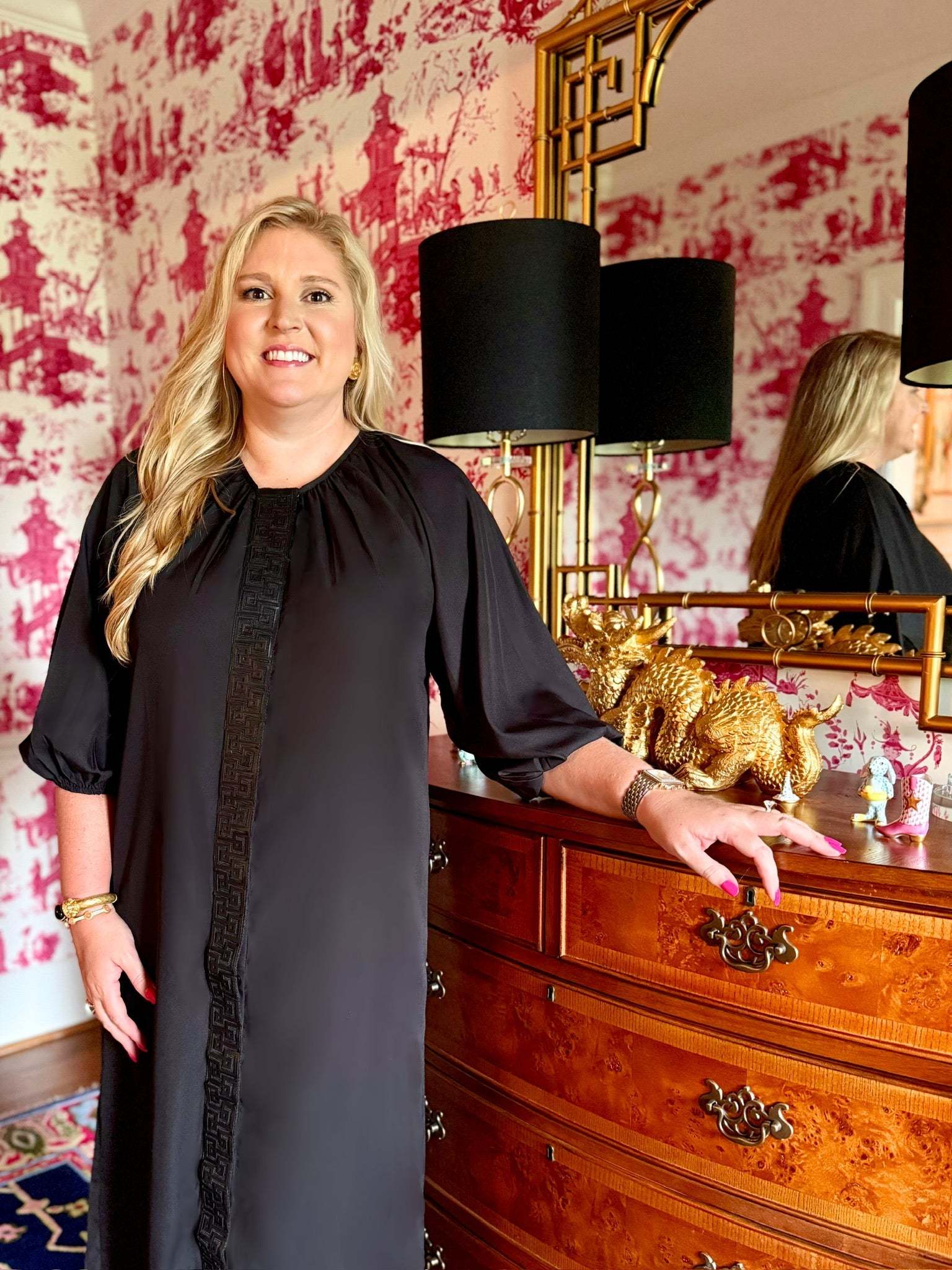 Woman wearing black Fall Hampton Dress with leather Greek key detail standing by wooden dresser with gold dragon decor