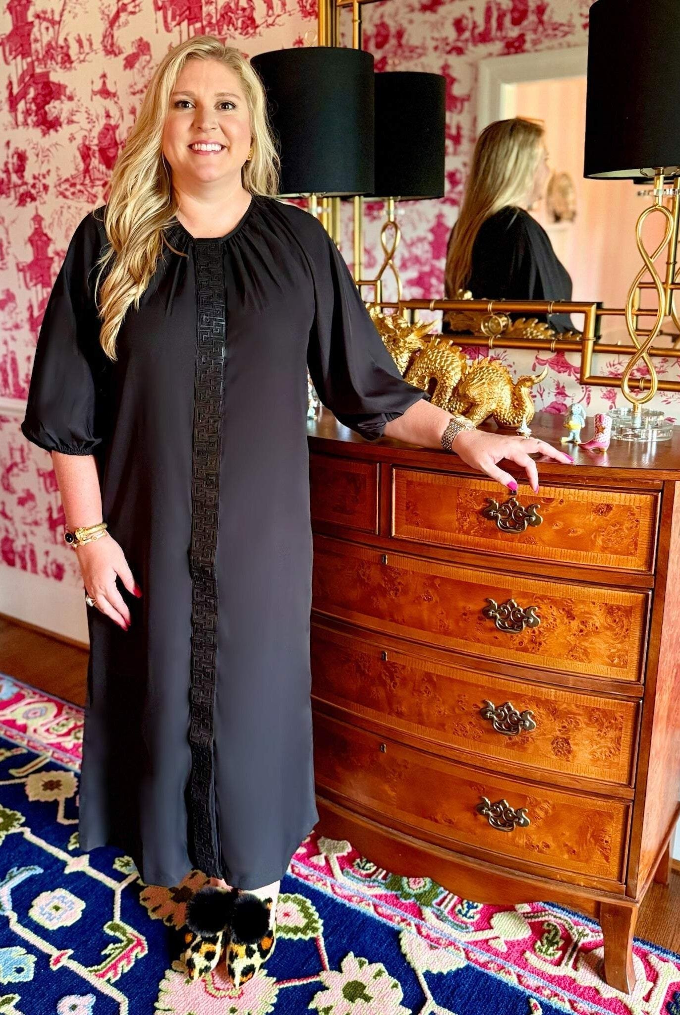 Woman wearing sleek black Fall Hampton Dress with leather Greek key detail on front placket standing next to wooden dresser in stylish room
