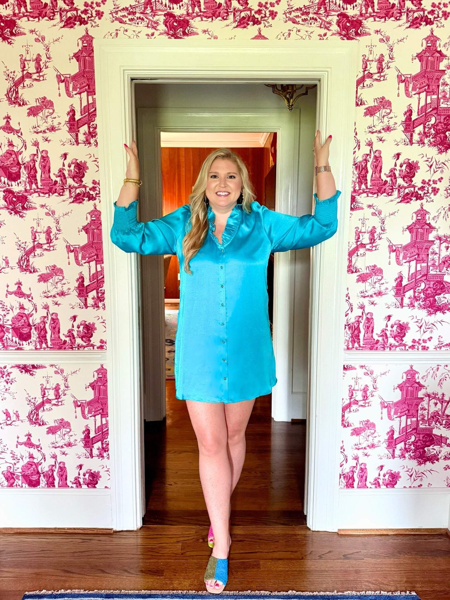 Woman wearing Harper Long Sleeve Dress in blue with ruffle neckline and gold buttons standing in doorway