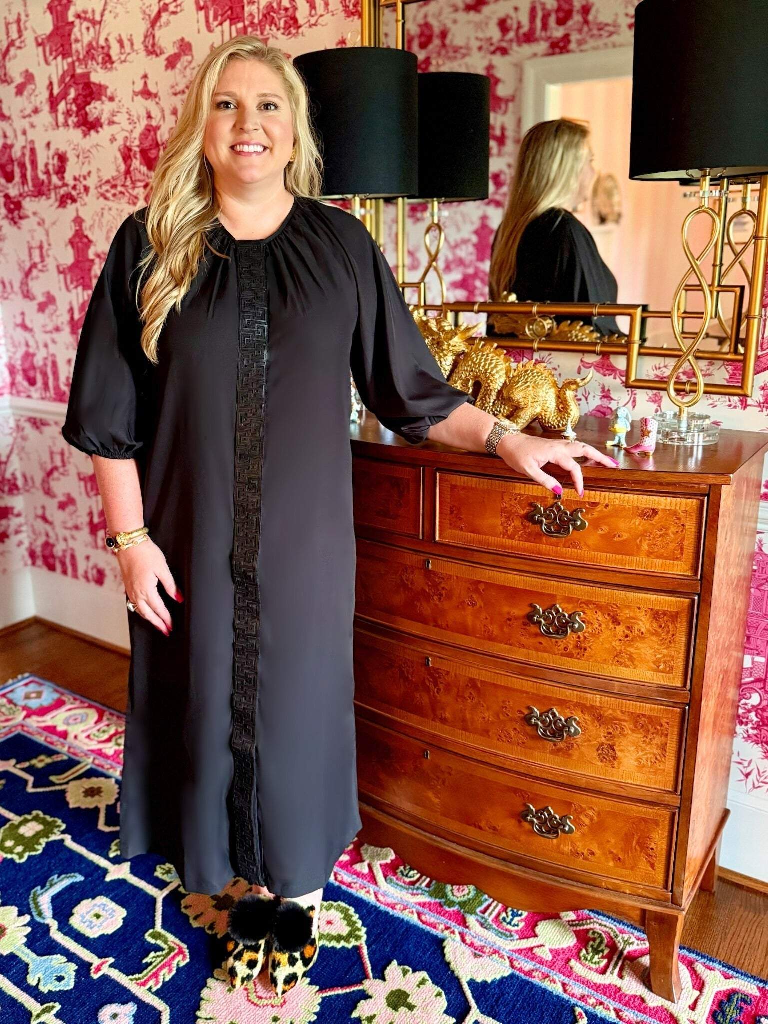Woman wearing sleek black Fall Hampton Dress with leather Greek key detail on front placket standing next to wooden dresser in stylish room